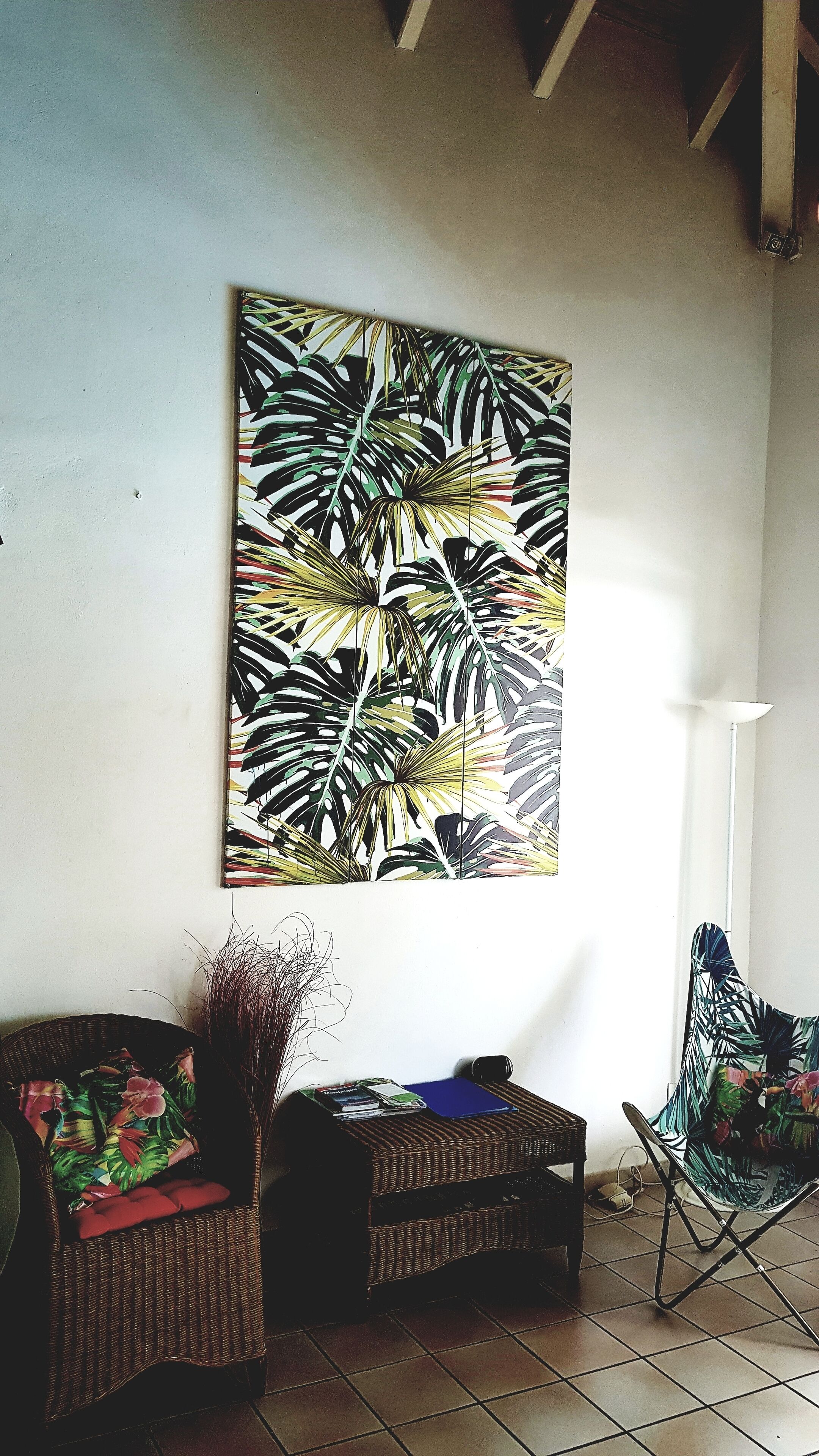 Une pièce décorée d’une grande œuvre d’art représentant des motifs de feuilles tropicales sur le mur, meublée de meubles en osier et agrémentée d’un lampadaire.