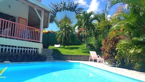 Una piscina con superficie de agua azul cristalina, rodeada de exuberante follaje y palmeras. Hay una silla de descanso blanca y una pequeña mesa blanca cerca de la piscina.
