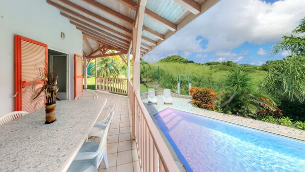 Un espacio exterior techado con piscina, sillas de descanso blancas y vista a un paisaje verde exuberante.
