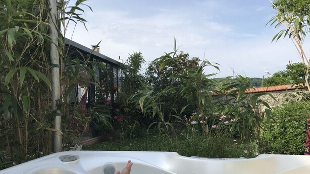 Een kuip met voeten ondergedompeld in water, omgeven door weelderige begroeiing en een huis op de achtergrond.