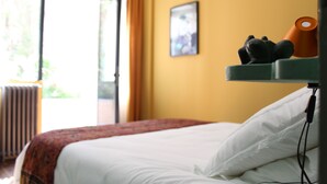 Een hotelkamer met een bed, een radiator, een raam met gordijnen, en een plank met een lamp en een decoratief object.