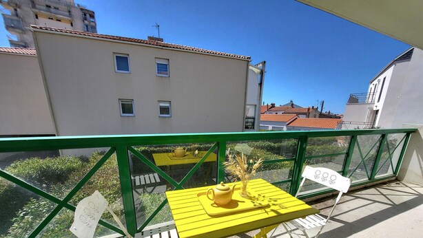 A balcony with a yellow table and white chairs, a green railing, and a view of buildings and trees. - Cozy T2 Apartment with South-Facing Balcony and Private Parking Near Beach (Les Sables-d'Olonne)