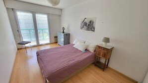 A bedroom with a bed, a dresser, a chair, and a window with a view. - Cozy T2 Apartment with South-Facing Balcony and Private Parking Near Beach (Les Sables-d'Olonne)