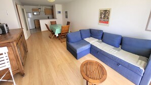 A living room with a blue sectional sofa, a wooden dining table with chairs, a small round wooden table, and a kitchen area in the background. - Cozy T2 Apartment with South-Facing Balcony and Private Parking Near Beach (Les Sables-d'Olonne)