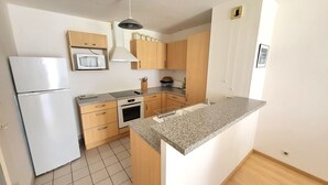 A compact kitchen with a refrigerator, microwave, stove, and wooden cabinets. - Cozy T2 Apartment with South-Facing Balcony and Private Parking Near Beach (Les Sables-d'Olonne)