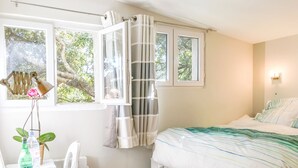 Une chambre avec un lit, une fenêtre avec des rideaux, une petite table sur laquelle se trouvent une bouteille et une plante, et un escalier menant au lit.