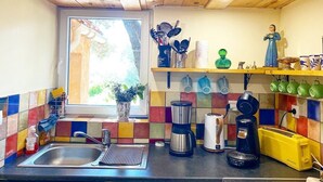 Une cuisine avec un plafond en bois, une fenêtre, un crédence colorée en carreaux, un évier, une machine à café, un grille-pain et une étagère contenant des ustensiles de cuisine et une figurine décorative.