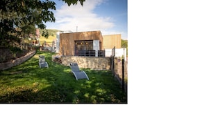 A modern outdoor patio with stone walls and a wooden deck, featuring two lounge chairs and a stone fireplace.
