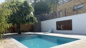 A swimming pool with a wooden deck, a lounge chair, and a stone wall with artwork.