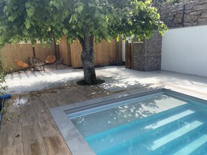 A backyard with a swimming pool, wooden deck, and a tree providing shade.
