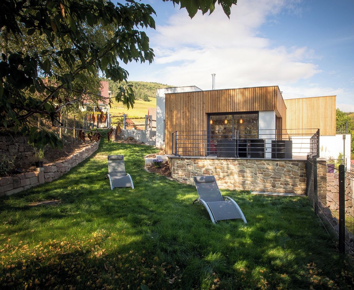 Een moderne, overdekte patio met stenen muren en een houten terras, met twee lounge stoelen en een stenen open haard.