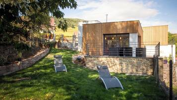 Een moderne, overdekte patio met stenen muren en een houten terras, met twee lounge stoelen en een stenen open haard.