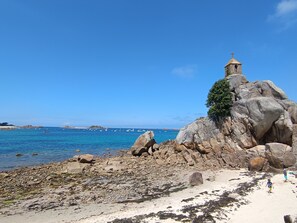 Een rotsachtig strand met een vuurtoren, helderblauwe zee en boten in de verte.