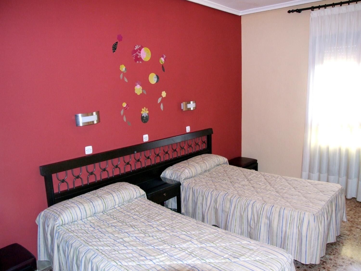 Habitación de hotel con dos camas, una pared roja y una ventana con cortinas blancas.