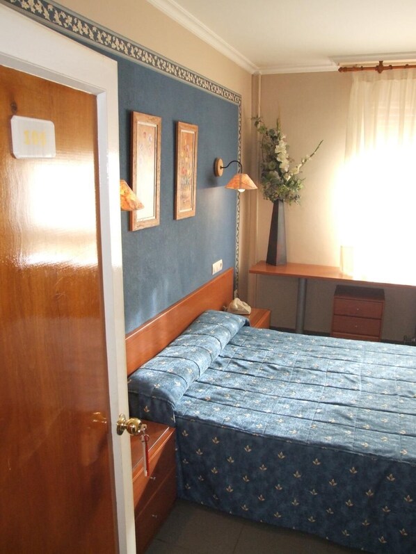 Quarto com porta de madeira, cama com colcha florida azul, duas fotos emolduradas na parede, mesa de cabeceira com abajur e janela com cortinas.
