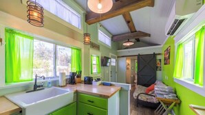 Een compacte keuken met een spoelbak, groene kastjes en een houten aanrecht. In de aangrenzende kamer staat een bed met een kleurrijk dekbed en kussens.