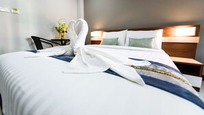 A neatly made bed with a towel arranged in a heart shape, a bedside table with a vase of flowers, and a headboard with two pillows.