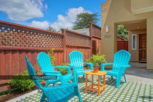 A patio with four turquoise Adirondack chairs and a small orange table, surrounded by a wooden fence and greenery.