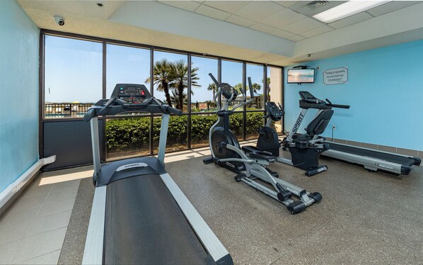 Un gimnasio con cintas de correr, una elíptica y una bicicleta estática, con vistas a palmeras y una piscina al aire libre.