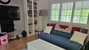 A living room with a blue sofa, white coffee table, bookshelf, television, and a window with a view of trees. - Private cozy cottage on Bobs Lake with boat launch (Maberly)