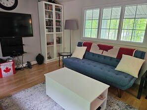 A living room with a blue sofa, white coffee table, bookshelf, television, and a window with a view of trees.