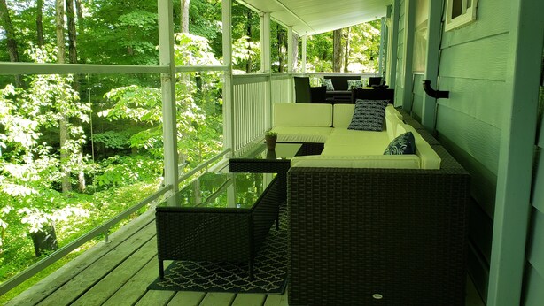 A covered porch with wicker furniture, a glass table, and a view of a lush green forest. - Private cozy cottage on Bobs Lake with boat launch (Maberly)