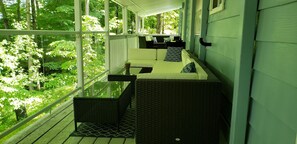 A covered porch with wicker furniture, a glass table, and a view of a lush green forest.