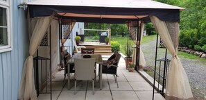 Ein überdachter Patio-Bereich mit Tisch und Stühlen, ein kleiner Garten und Blick ins umliegende Grün.