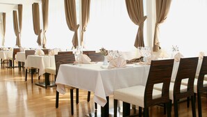 Un comedor con mesas dispuestas para el servicio, sillas de madera y ventanas grandes con cortinas.
