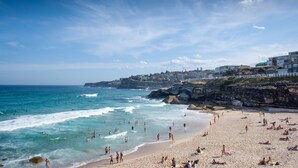 Une plage où l’on voit des gens nager et bronzer, une côte rocheuse et des bâtiments en arrière-plan.