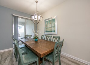 A dining room with a wooden table, six chairs, a vase with flowers, a chandelier, and a painting on the wall.