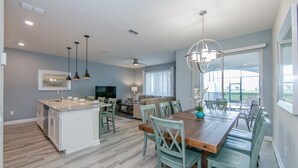 A modern kitchen with a central island, dining area with a wooden table and chairs, and a living area with a sofa and TV. - (JA4907)  Home 4/3 Private Pool, Lake & Free Waterpark (Kissimmee)