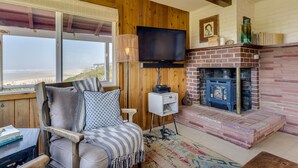 A cozy living room with a wooden paneled wall, a television, a brick fireplace, and a view of the beach.