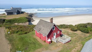 A red house with a porch, located near a beach with a rocky shoreline and a lighthouse in the background.