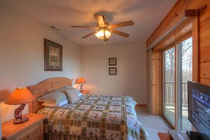 Une chambre avec un ventilateur de plafond en bois, un lit recouvert d’une courtepointe, deux lampes de chevet et un téléviseur posé sur un meuble en bois.