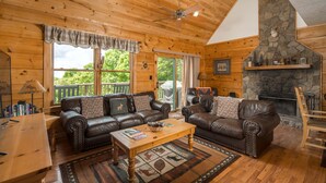 A log cabin interior with two leather sofas, a wooden coffee table, a stone fireplace, and a balcony with a view of trees. - Sleeps 10 | Hot Tub, Game Room, Mountain Views & Near Beech Ski Resort (Beech Mountain)
