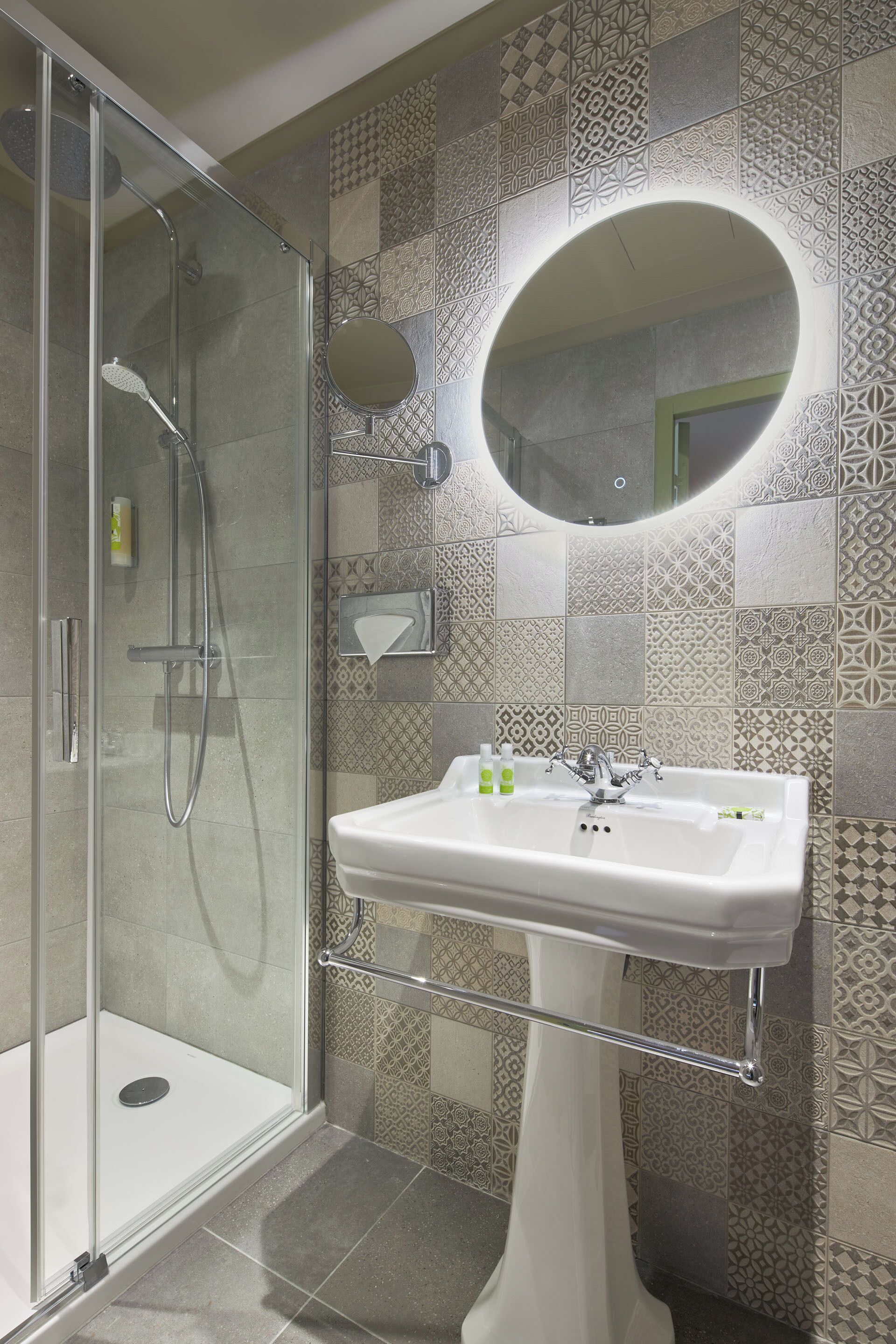 A modern bathroom with a glass-enclosed shower, a pedestal sink, and patterned tiles.