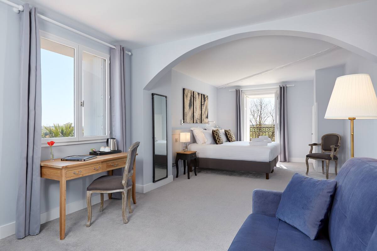 A hotel room with a bed, a desk, a chair, a sofa, a lamp, and a window with curtains.