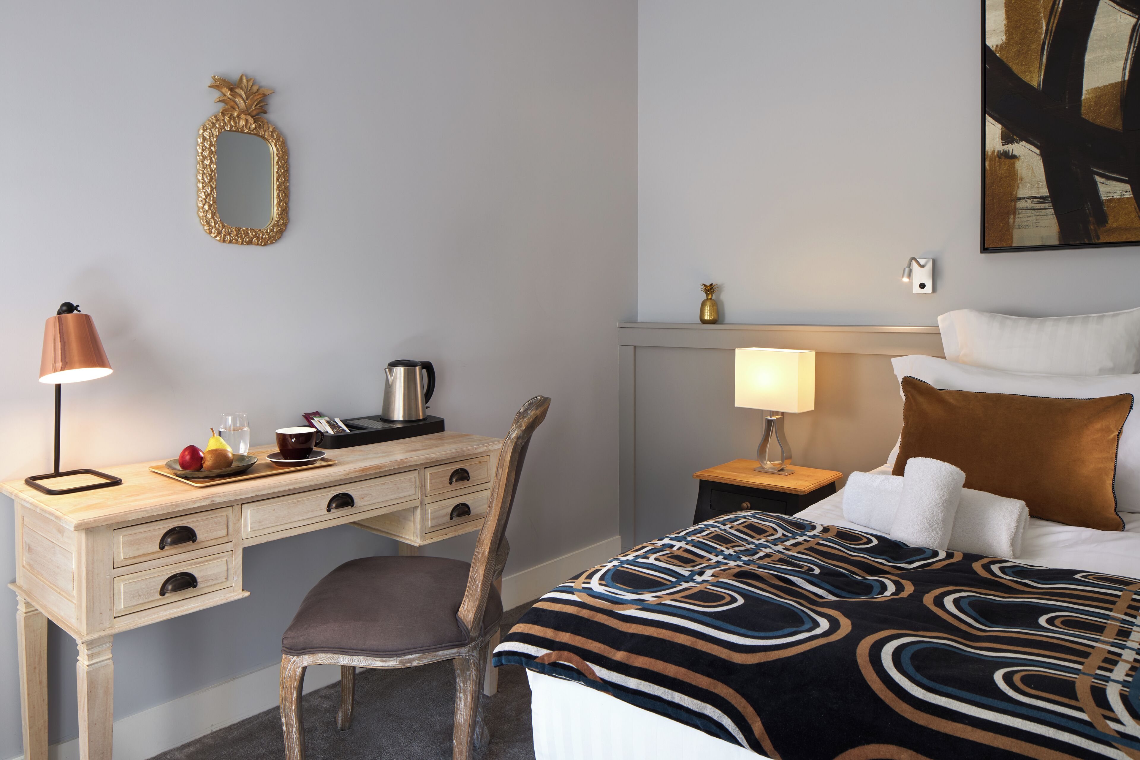 A hotel room with a bed, a desk with a lamp, a chair, a nightstand with a lamp, and a wall-mounted mirror.