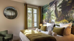 Quarto de hotel com cama, uma cadeira, janela com cortinas e um grande mural com tema de floresta na parede.
