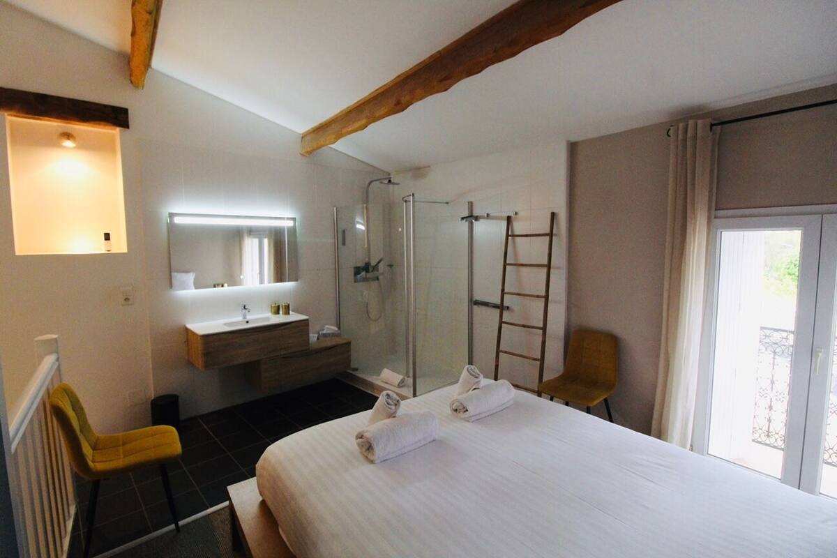 A hotel room with a large bed, a bathroom with a glass shower, a wooden ceiling, and a view of the outdoors.