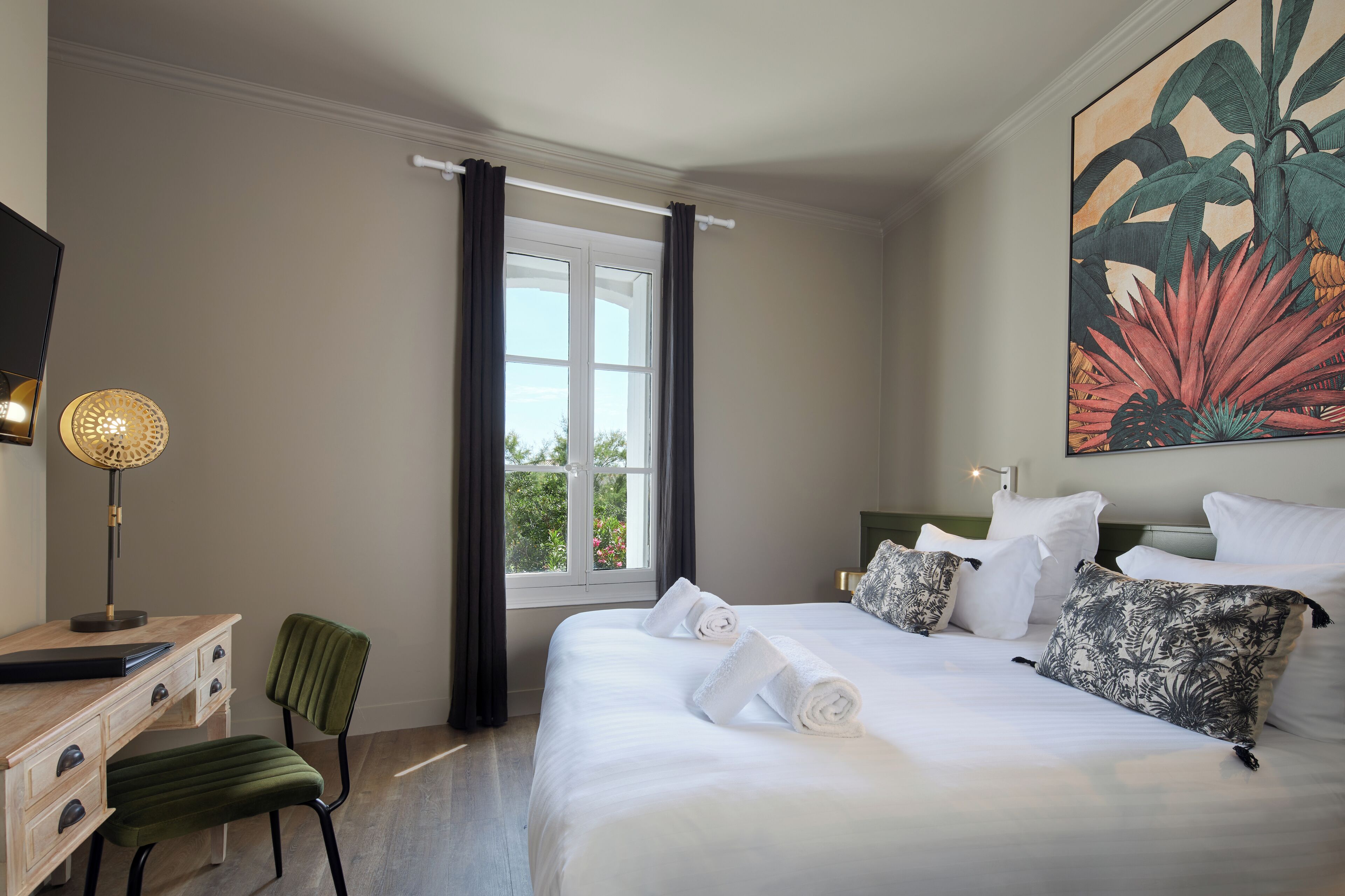 A hotel room with a bed, a desk, a chair, a window with curtains, and a painting on the wall.