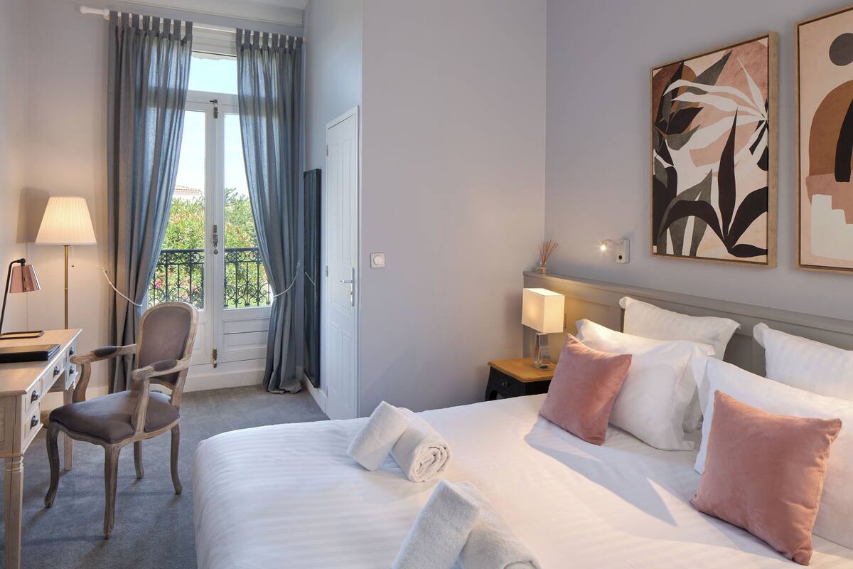 A hotel room with a bed, a desk, a chair, a nightstand, and artwork on the wall.