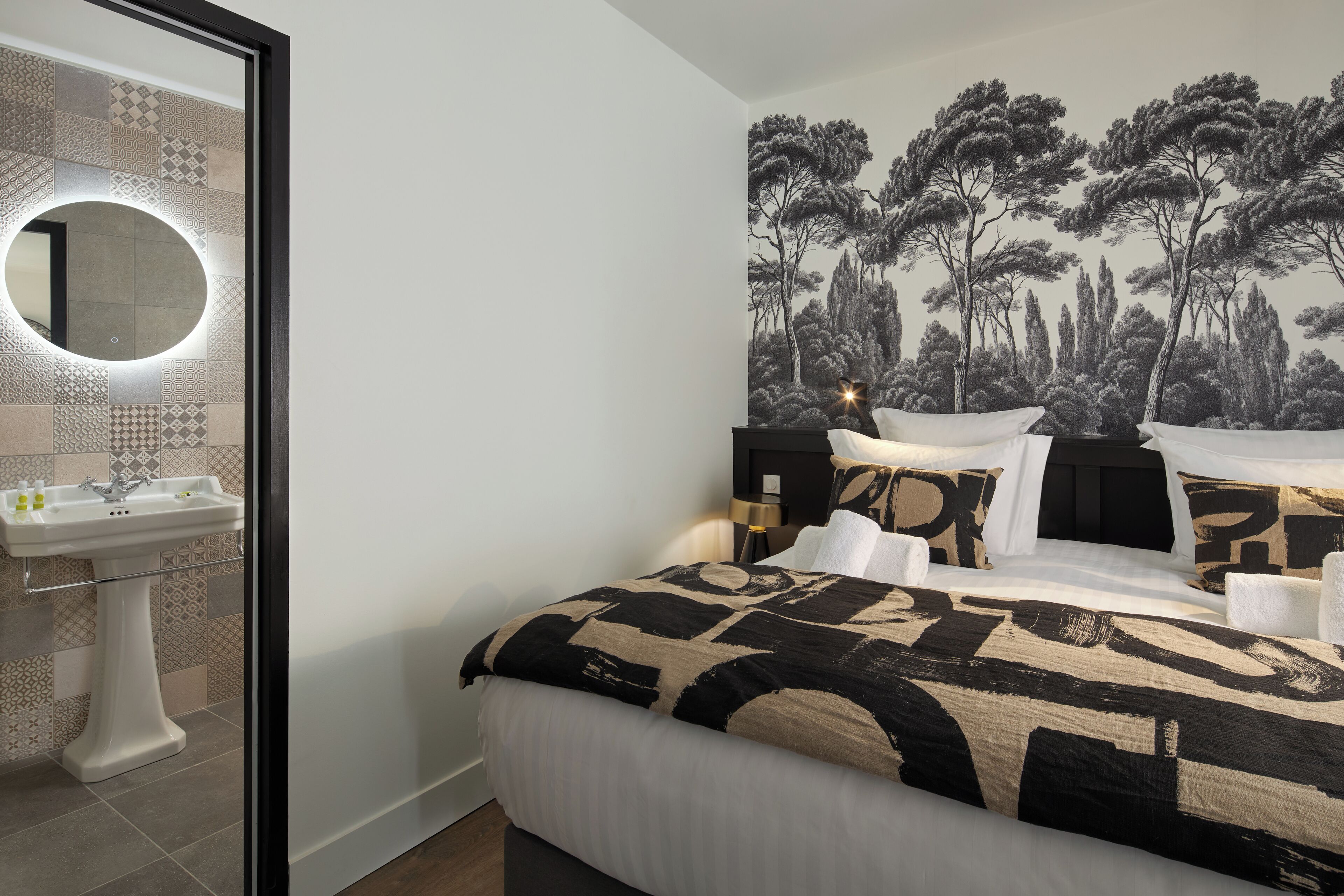 A bedroom with a bed, a nightstand, a wall-mounted mirror, and a bathroom with a sink and tiles.