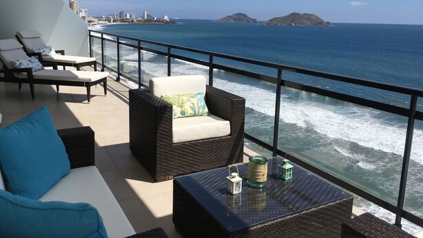 A balcony with wicker furniture, a glass table with decorative items, and a view of the ocean and distant cityscape.