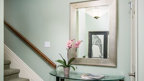 Un escalier avec une rampe en bois, une console en verre sur laquelle sont posés un pot de plantes et des magazines, et une œuvre d’art encadrée représentant un éléphant, accrochée au mur.