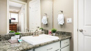 Une salle de bain avec un lavabo blanc, un comptoir en granit, un miroir, deux crochets pour serviettes, une plante en pot et un petit meuble.