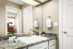 Une salle de bain avec un lavabo blanc, un comptoir en granit et un miroir. Des serviettes sont accrochées à des crochets et une petite plante en pot est posée sur le comptoir.