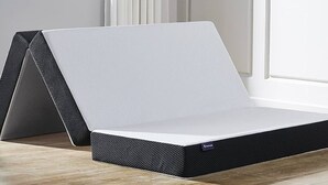 Una cama moderna y minimalista con colchón blanco y estructura negra, ubicada en una habitación con suelo de madera clara y paredes blancas.