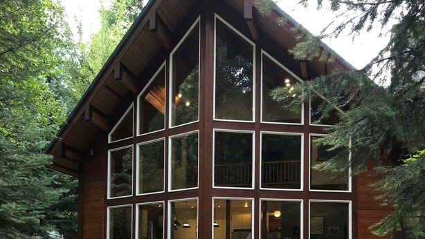 A wooden cabin with large windows, a stone foundation, and outdoor seating.